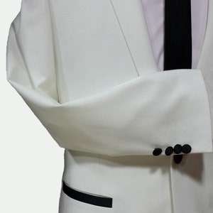 May include: A white tuxedo jacket with a black satin lapel and black buttons. The jacket has a black pocket trim and a single button closure.
