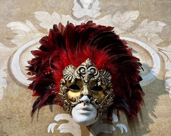 May include: A gold and white masquerade mask with red feathers. The mask has intricate details and a feminine design.