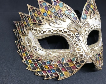 May include: A gold masquerade mask with a colorful checkered pattern and intricate details. The mask has a single eye opening and is decorated with glitter.