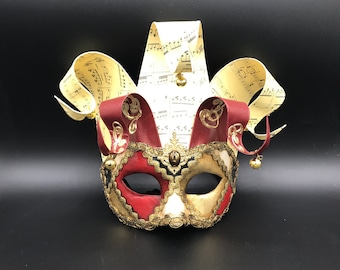 May include: A red, gold, and white masquerade mask with a jester's hat. The hat is made of paper with musical notes printed on it. The mask has a black eye mask and gold accents.