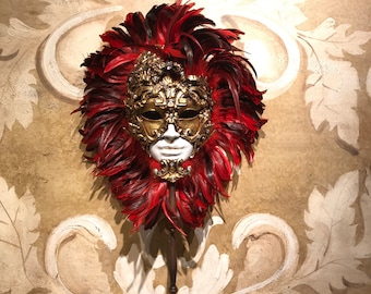 May include: A white and gold Venetian masquerade mask with red feathers. The mask is decorated with intricate details and is mounted on a wooden stick.