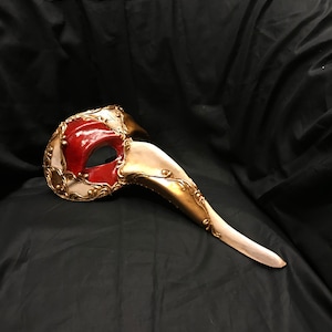 Peut inclure: Un masque de carnaval blanc et or avec un long nez incurvé et des accents rouges. Le masque est décoré de détails complexes et a un style vénitien traditionnel.