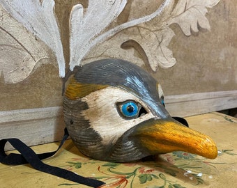 May include: A hand-painted bird mask with a black strap. The mask features a gray and white head with a yellow beak and bright blue eyes. The mask is resting on a floral patterned surface.