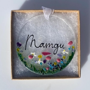 May include: A round glass bauble with a floral design and the word "Mamgu" written in black script. The bauble is in a brown box with white tissue paper.