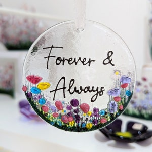 Op de afbeelding: Een transparante glazen ornament met een wit lint, met een kleurrijk bloemenontwerp en de woorden "Forever & Always" in het zwart.