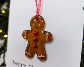 May include: A clear, gingerbread man ornament with red button details and a red ribbon for hanging. The ornament is hanging in front of a white card with the words "Merry Christmas" printed in black.