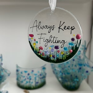 Może przedstawiać: Okrągła szklana ozdoba z białym tłem i zieloną obwódką. Ozdoba ma kwiatowy wzór z niebieskimi, różowymi i żółtymi kwiatami. Tekst "Always Keep Fighting" jest napisany czarnym kursywą na ozdobie.