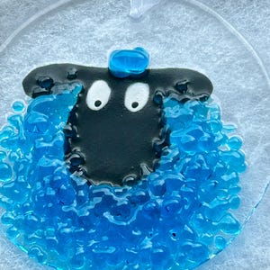 May include: A blue glass sheep ornament with a black face and white eyes. The sheep has a blue glass hat on its head.