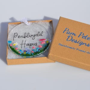 Puede incluir: Un adorno de vidrio transparente y redondo con las palabras "Penblwydd Hapus" y un diseño floral, presentado en una pequeña caja de cartón. La caja está abierta, revelando el adorno. La caja tiene el texto "Pam Peters Designs" en azul.