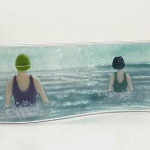 May include: A curved glass art piece depicting two figures in swimsuits and swim caps in the ocean. One figure wears a purple swimsuit and green cap, the other a green swimsuit and black cap. The water is a mix of blue and white.