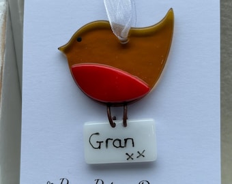 May include: A glass robin ornament with a red breast and brown wings. The ornament has a white tag with the word "Gran" written on it in black marker. The tag also has two black "x" marks. The ornament is hanging from a white ribbon. The background is white with a black floral design and the text "Pam Peters Designs".
