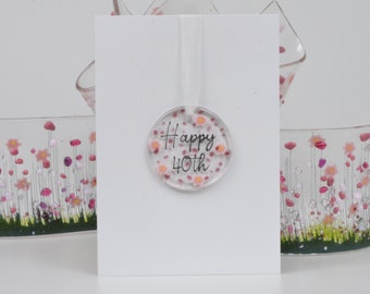 May include: A white card with the text "Happy 40th" on a clear round glass ornament with pink and white flowers. The ornament is hanging from a white ribbon.