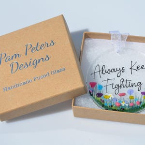 May include: A clear glass ornament with the words "Always Keep Fighting" and a colorful floral design. The ornament is in a brown box with the text "Pam Peters Designs" and "Handmade Fused Glass".