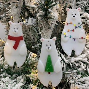 May include: Three white polar bear Christmas tree decorations. One has a red scarf, another a green tree, and the third is strung with colourful lights. Each ornament is suspended by a sheer ribbon, against a backdrop of a snow-covered Christmas tree.