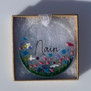 May include: A round glass bauble with a floral design and the name "Nain" written in black ink. The bauble is surrounded by a white background.