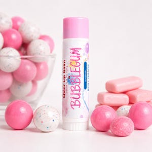 May include: A cylindrical lip balm tube with a pink cap and white label featuring the text Sheer Lip Balm in BUBBLEGUM, Net Wt. 0.15 oz / 4.25 g, and Roxy Grace. The tube is surrounded by pink and white speckled gumballs and rectangular gum pieces.