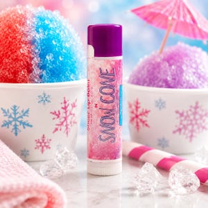 Puede incluir: Un tubo cil&iacute;ndrico de b&aacute;lsamo labial con tapa morada y una etiqueta con un patr&oacute;n de m&aacute;rmol rosa y blanco. El texto en la etiqueta dice Sheer Lip Balm in SNOW COVE con un peso neto de 4,25 g. El tubo est&aacute; colocado en posici&oacute;n vertical.
