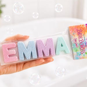 May include: A set of four letter-shaped bath bombs spelling the name EMMA in pink, blue, purple, and light green, packaged in a clear plastic sleeve with a colorful tie-dye patterned label that reads BATH BOMBS and Play Grace.