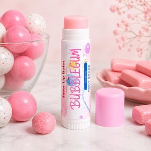 May include: A white lip balm tube with a pink cap, labeled Sheer Lip Balm in BUBBLEGUM by ROXY GRAGE, net weight 0.15 oz, 4.25 g. The tube is surrounded by pink and white speckled gumballs in a glass bowl and pink rectangular gum pieces.