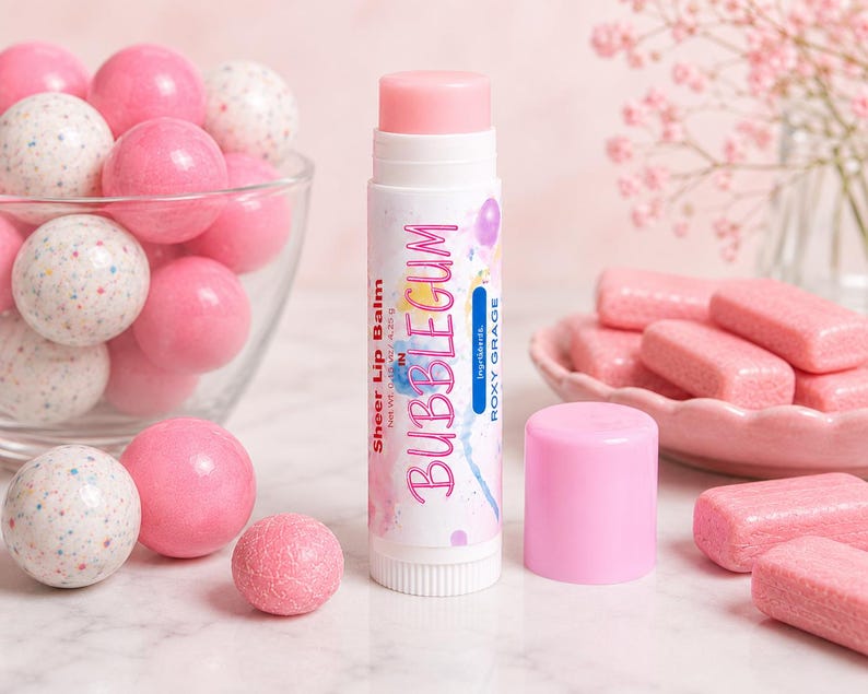 May include: A white lip balm tube with a pink cap, labeled Sheer Lip Balm in BUBBLEGUM by ROXY GRAGE, net weight 0.15 oz, 4.25 g. The tube is surrounded by pink and white speckled gumballs in a glass bowl and pink rectangular gum pieces.
