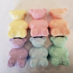 Six colorful bear-shaped bath bombs in yellow, pink, peach, purple, blue, and green are arranged in two rows on a light surface.