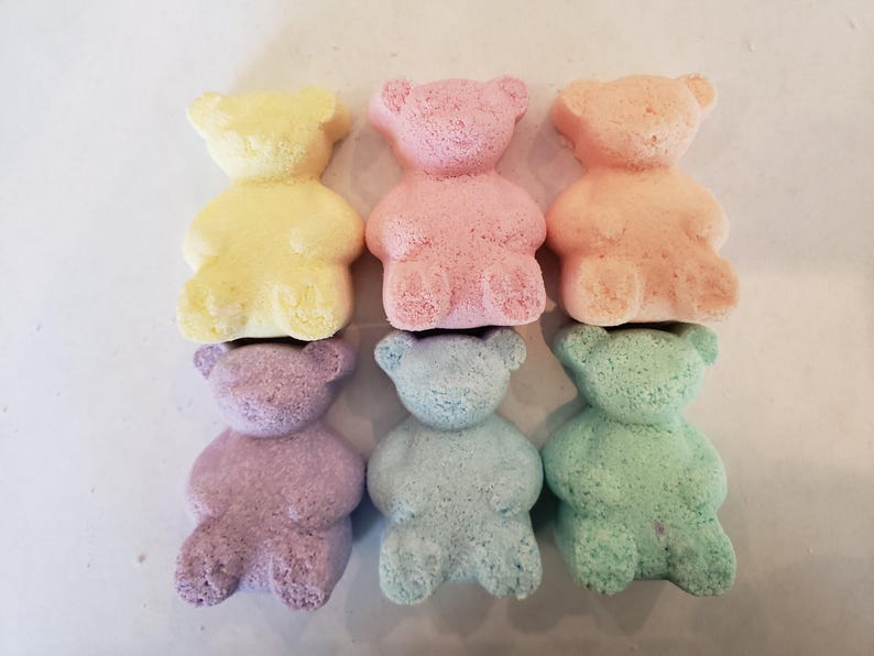 Six colorful bear-shaped bath bombs in yellow, pink, peach, purple, blue, and green are arranged in two rows on a light surface.