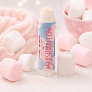 May include: A cylindrical lip balm tube with a light blue and pink cloud-patterned label. The text reads Sheer Lip Balm in MARSHMALLOW, Net Wt. 0.15 oz / 4.25 g. The tube has a white cap and is surrounded by several white and pink marshmallows.