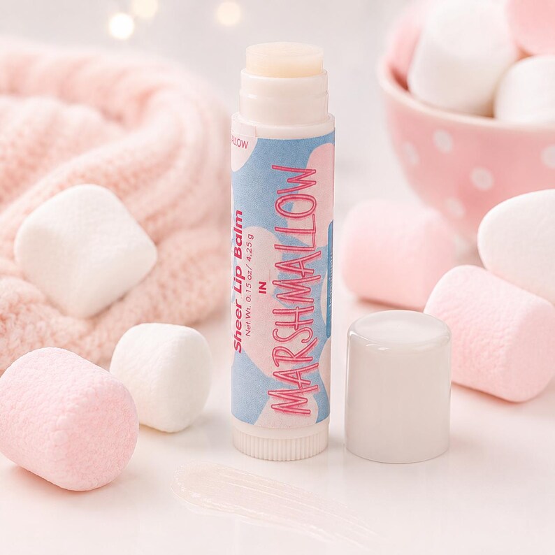 May include: A cylindrical lip balm tube with a light blue and pink cloud-patterned label. The text reads Sheer Lip Balm in MARSHMALLOW, Net Wt. 0.15 oz / 4.25 g. The tube has a white cap and is surrounded by several white and pink marshmallows.