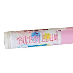 May include: A cylindrical lip balm tube with a light pink cap and a white label featuring watercolor splashes in blue, yellow, and pink. The text reads Sheer Lip Balm, Net Wt. 0.15 oz / 4.25 g, IN BUBBLEGUM, and Ingredients. The tube has a white twist base.