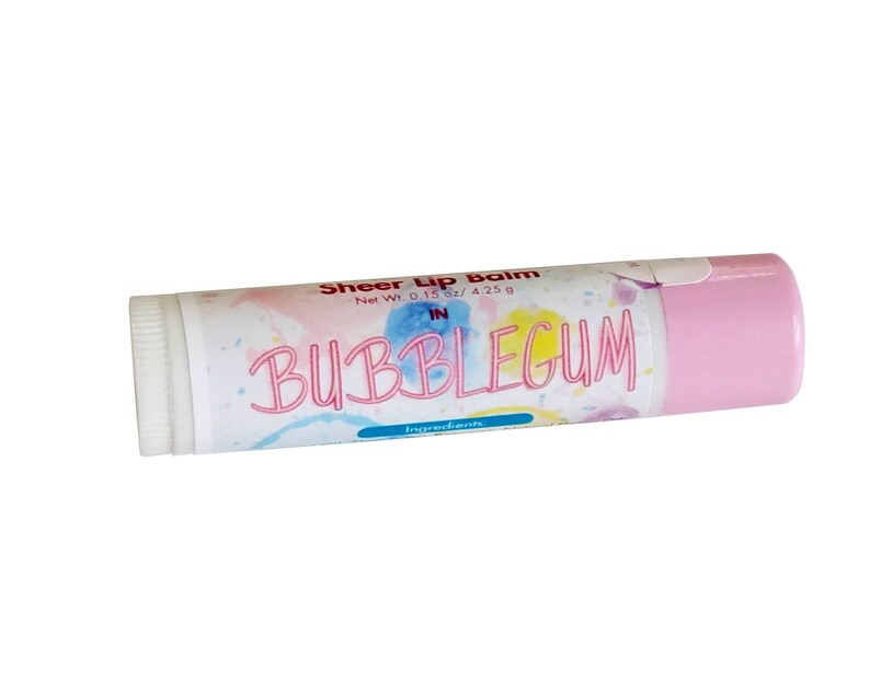 May include: A cylindrical lip balm tube with a light pink cap and a white label featuring watercolor splashes in blue, yellow, and pink. The text reads Sheer Lip Balm, Net Wt. 0.15 oz / 4.25 g, IN BUBBLEGUM, and Ingredients. The tube has a white twist base.
