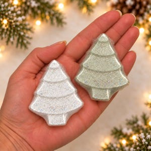 May include: Two Christmas tree-shaped bath bombs wrapped in clear plastic. One bath bomb is white with a fine glitter finish, and the other is a pale sage green with a similar sparkling, glittery texture. Both items feature a tiered design.