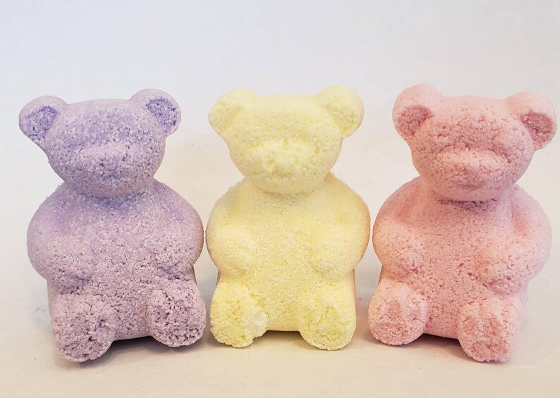 Three bear-shaped bath bombs in purple, yellow, and pink stand in a row against a white background.