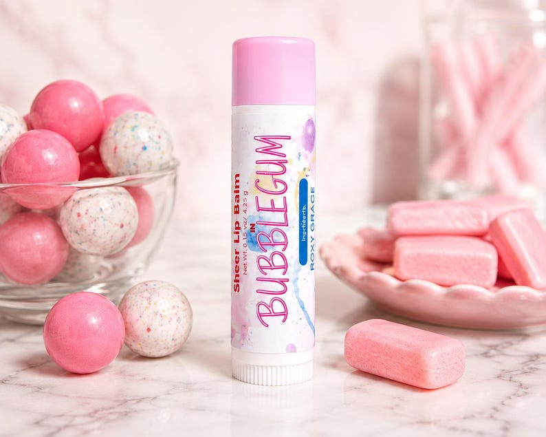 May include: A cylindrical lip balm tube with a pink cap and white label featuring the text Sheer Lip Balm, BUBBLEGUM, and Roxy Grace. The label includes watercolor accents and states a net weight of 0.15 oz / 4.25 g.