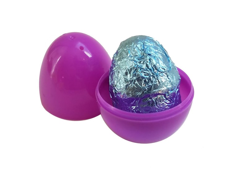 May include: A purple plastic egg, opened to reveal a foil-wrapped bath bomb egg. The foil has blue and silver tones. The other half of the egg is next to the open half. The background is white.