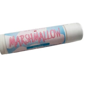 May include: A cylindrical lip balm tube with a white cap and a light blue label featuring a white cloud pattern. The text MARSHMALLOW is printed in large pink letters across the center of the label, with smaller text listing ingredients below.