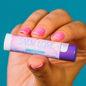 Puede incluir: Un tubo cil&iacute;ndrico de b&aacute;lsamo labial con tapa blanca y base morada. La etiqueta presenta un patr&oacute;n de nubes rosas y azules con el texto SNOW CONE en letra blanca. Debajo del texto, la palabra Ingredients est&aacute; impresa sobre una lista de componentes.