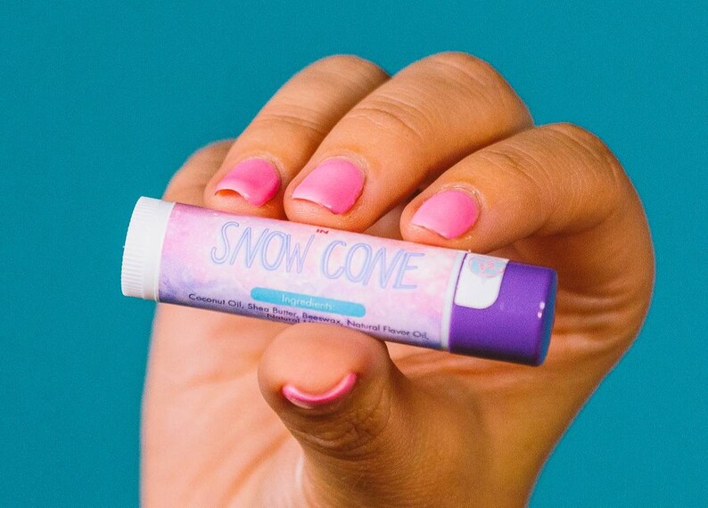 Puede incluir: Un tubo cil&iacute;ndrico de b&aacute;lsamo labial con tapa blanca y base morada. La etiqueta presenta un patr&oacute;n de nubes rosas y azules con el texto SNOW CONE en letra blanca. Debajo del texto, la palabra Ingredients est&aacute; impresa sobre una lista de componentes.