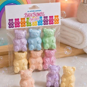 A package of six bear bath bombs stands upright behind four loose bears near a candle and a rainbow prop.