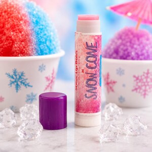 Puede incluir: Un tubo cil&iacute;ndrico de b&aacute;lsamo labial con una etiqueta jaspeada en rosa y blanco que incluye el texto "Sheer Lip Balm IN SNOW CONE" y "Net Wt. 0.15 oz / 4.25 g". El tubo tiene una punta de b&aacute;lsamo rosa claro y una tapa morada s&oacute;lida al lado.