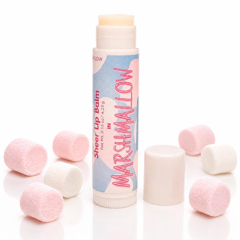May include: A cylindrical tube of sheer lip balm with a light pink and blue abstract pattern on the label. The text reads MARSHMALLOW, Sheer Lip Balm, and Net Wt. 0.15 oz / 4.25 g. The tube is surrounded by several small, soft pink and white marshmallows.