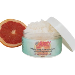 Könnte beinhalten: Ein weißes Glas mit einem hellblauen Etikett und einem rosafarbenen Hashtag, der #JUICY lautet. Das Etikett sagt auch "Find your happy place and recharge with uplifting grapefruit" und "All Natural Body Scrub 8 fl oz". Das Glas ist offen und eine Grapefruit-Hälfte liegt daneben.
