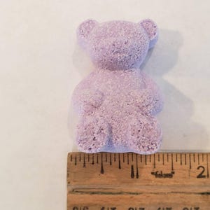 A single purple bear-shaped bath bomb rests above a wooden ruler showing its size is about two inches tall.
