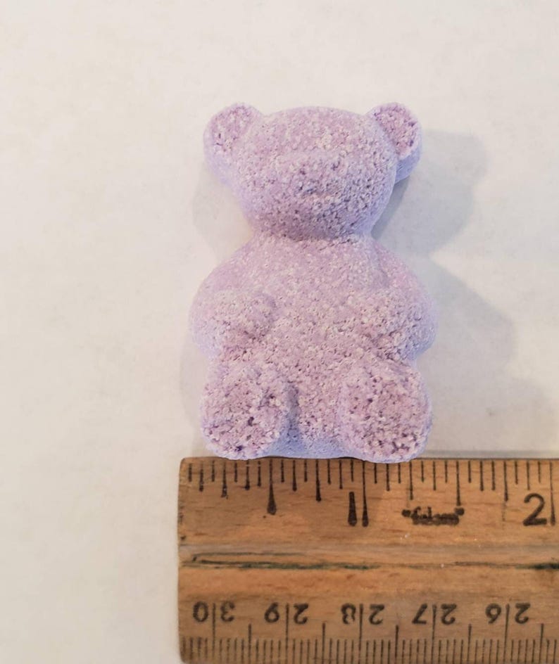 A single purple bear-shaped bath bomb rests above a wooden ruler showing its size is about two inches tall.