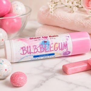 May include: A cylindrical tube of Roxy Grace sheer lip balm in Bubblegum flavor, labeled with a net weight of 0.15 oz / 4.25 g. The white label features pink and blue watercolor accents, surrounded by pink gumballs and rectangular gum pieces.