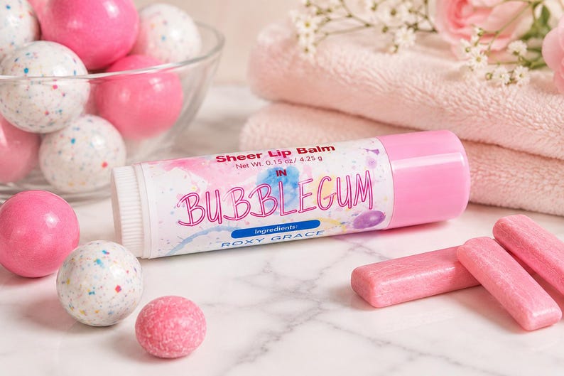 May include: A cylindrical tube of Roxy Grace sheer lip balm in Bubblegum flavor, labeled with a net weight of 0.15 oz / 4.25 g. The white label features pink and blue watercolor accents, surrounded by pink gumballs and rectangular gum pieces.