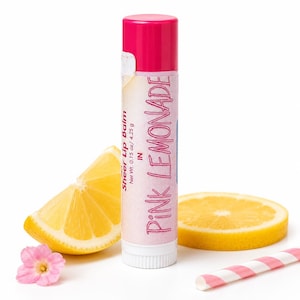 Könnte beinhalten: Ein zylindrischer Lippenbalsamstift mit einer leuchtend pinken Kappe und einem Etikett mit der Aufschrift Sheer Lip Balm in Pink Lemonade, Net Wt. 0.15 oz / 4.25 g. Der Stift ist umgeben von einem gelben Zitronenschnitz, einer Zitronenscheibe, einer kleinen rosa Blume und einem gestreiften Strohhalm.