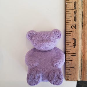 A purple bear-shaped bath bomb sits next to a wooden ruler measuring slightly over two inches tall.