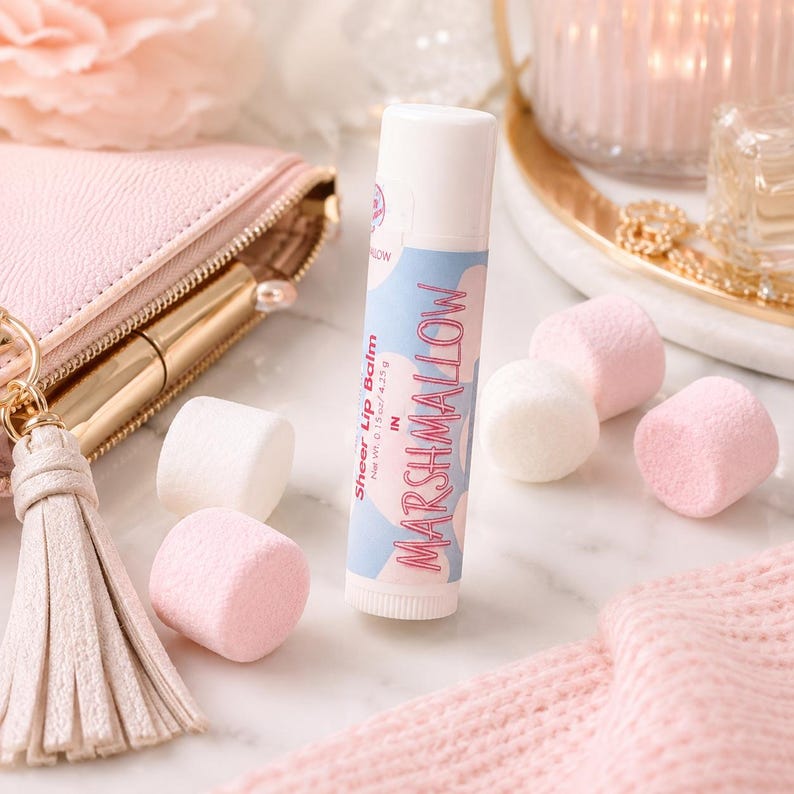 May include: A cylindrical lip balm tube with a white cap and a label featuring light blue and pink cloud shapes. The text on the label reads Sheer Lip Balm in MARSHMALLOW with a net weight of 0.15 oz / 4.25 g printed clearly on the side.