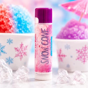 Puede incluir: Un tubo cil&iacute;ndrico de b&aacute;lsamo labial con tapa de color morado oscuro y base blanca. La etiqueta presenta un fondo texturizado en rosa y blanco con el texto Sheer Lip Balm IN SNOW CONE y Net Wt. 0.15 oz/4.25 g en letras moradas.