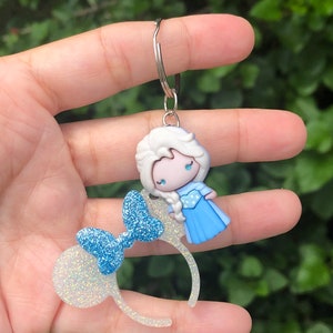 May include: A white and blue glitter Minnie Mouse ear headband keychain with a small blue glitter bow and a blue and white character charm attached.
